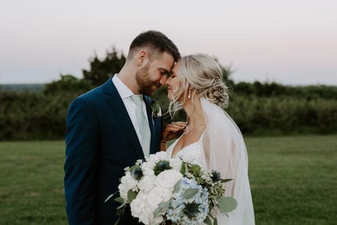 Groom and bride touching foreheads at dusk, bride holding a white and blue bouquet